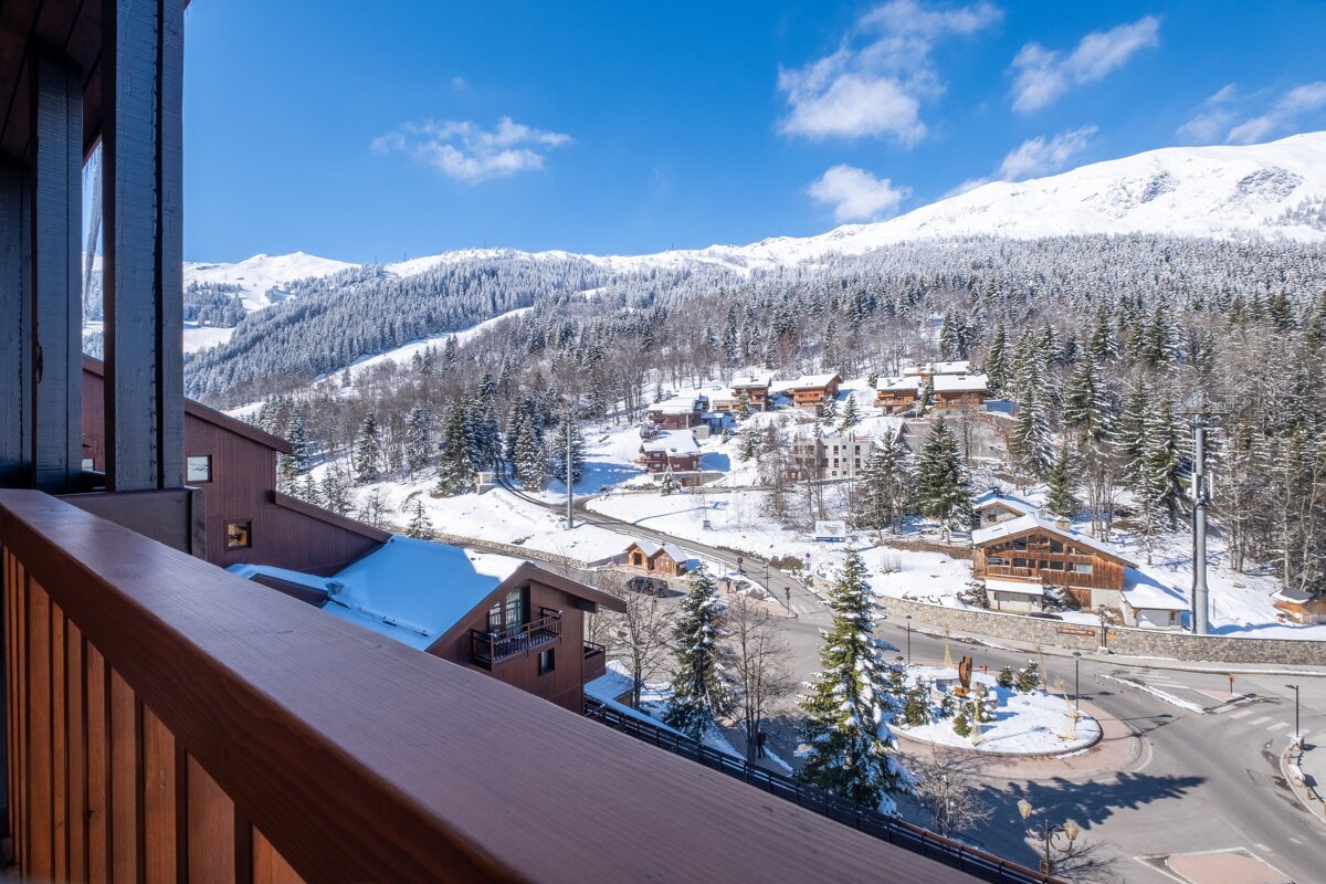 A stunning balcony view of a snow-covered alpine village with chalets nestled in pine forests, backed by vast snowy mountains under a bright blue sky.