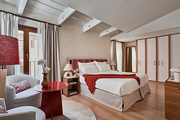 A stylish bedroom featuring a white bed with red accents, exposed beams, a balcony, and light wood flooring, creating a cozy and inviting space.