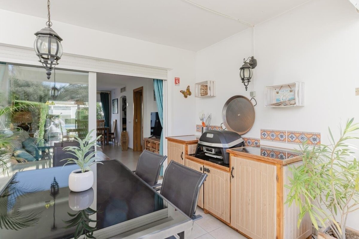 A bright patio dining area features a table, grill station with a tiled backsplash, potted plants, and sliding glass doors leading to an indoor room.