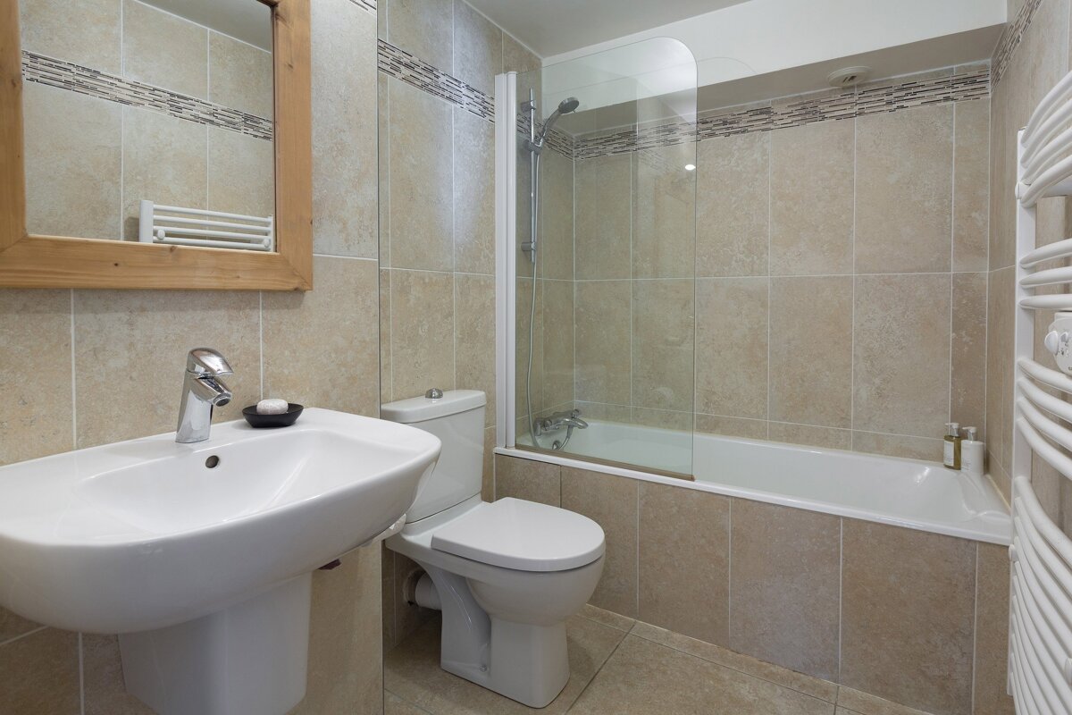 A modern bathroom with beige tiles, featuring a white sink, toilet, bathtub with shower screen, wooden mirror, and a white towel radiator.