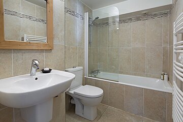 A modern bathroom with beige tiles, featuring a white sink, toilet, bathtub with shower screen, wooden mirror, and a white towel radiator.