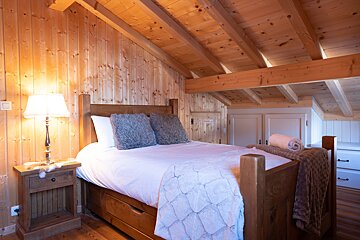 A cozy, rustic bedroom features warm wood paneling, exposed beams, a large bed with fluffy pillows, and a lit bedside lamp, creating an inviting ambiance.