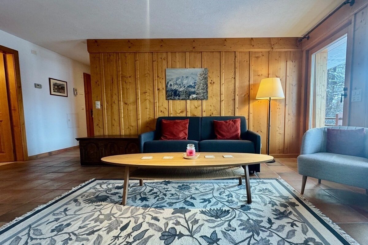 Inviting chalet-style living room with wood-paneled walls, blue sofa, oval coffee table, and a large window looking out to snow.