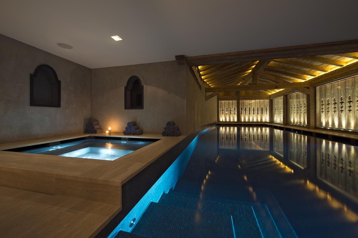 An elegant indoor pool and hot tub area with warm, ambient lighting, rustic wooden beams, and ornate carved panels, creating a tranquil and luxurious spa atmosphere.