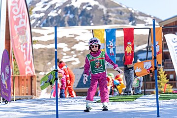 A young girl in pink ski gear, helmet, and sunglasses stands on a snowy slope in a kids' ski area with colorful banners and mountains behind.