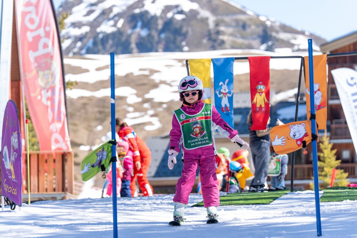 A young girl in a pink ski suit, helmet & sunglasses on a sunny ski slope, surrounded by colorful kids' ski school flags & distant mountains.