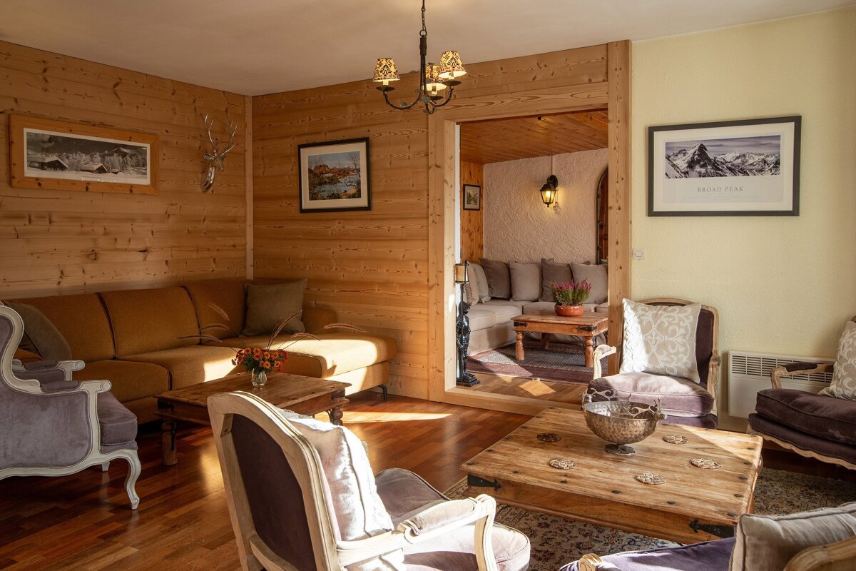 A warm, wood-paneled living room in a rustic chalet style. It features multiple seating areas, including sofas and armchairs, with an adjoining cozy room visible.