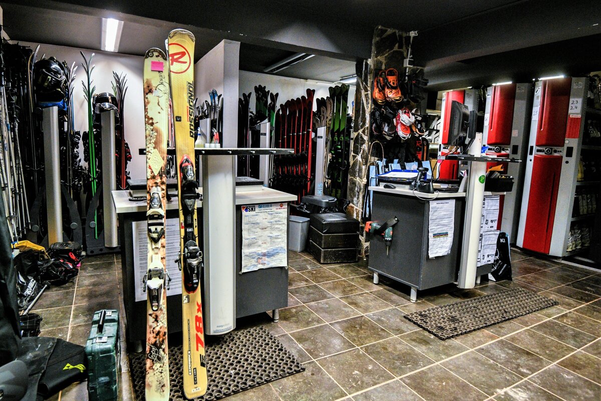 Interior of a dimly lit ski rental shop with rows of skis, boots, and counters. Two skis are prominently displayed in the foreground.