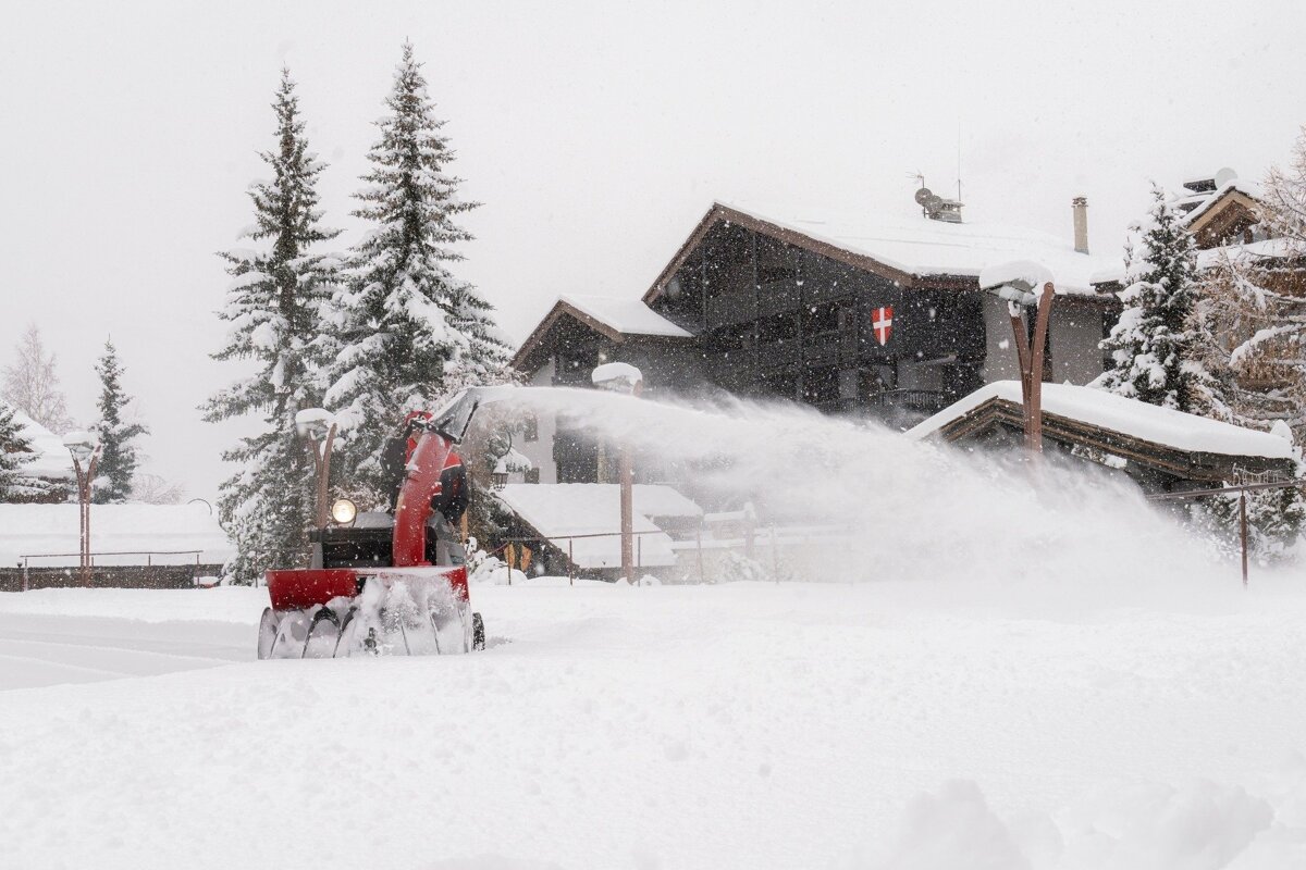 A red snowblower powerfully sprays snow, clearing a path in a heavily snow-covered village scene with buildings and pine trees under falling snow.