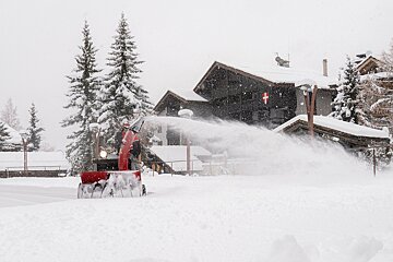 A red snowblower powerfully sprays snow, clearing a path in a heavily snow-covered village scene with buildings and pine trees under falling snow.