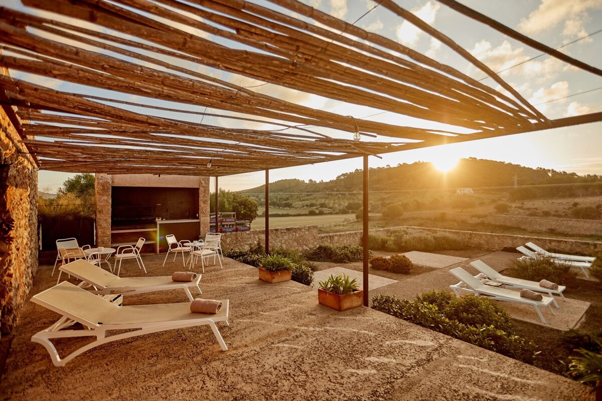 A rustic outdoor patio with a wooden pergola, sun loungers, and dining area, overlooking a beautiful rural landscape at sunset.