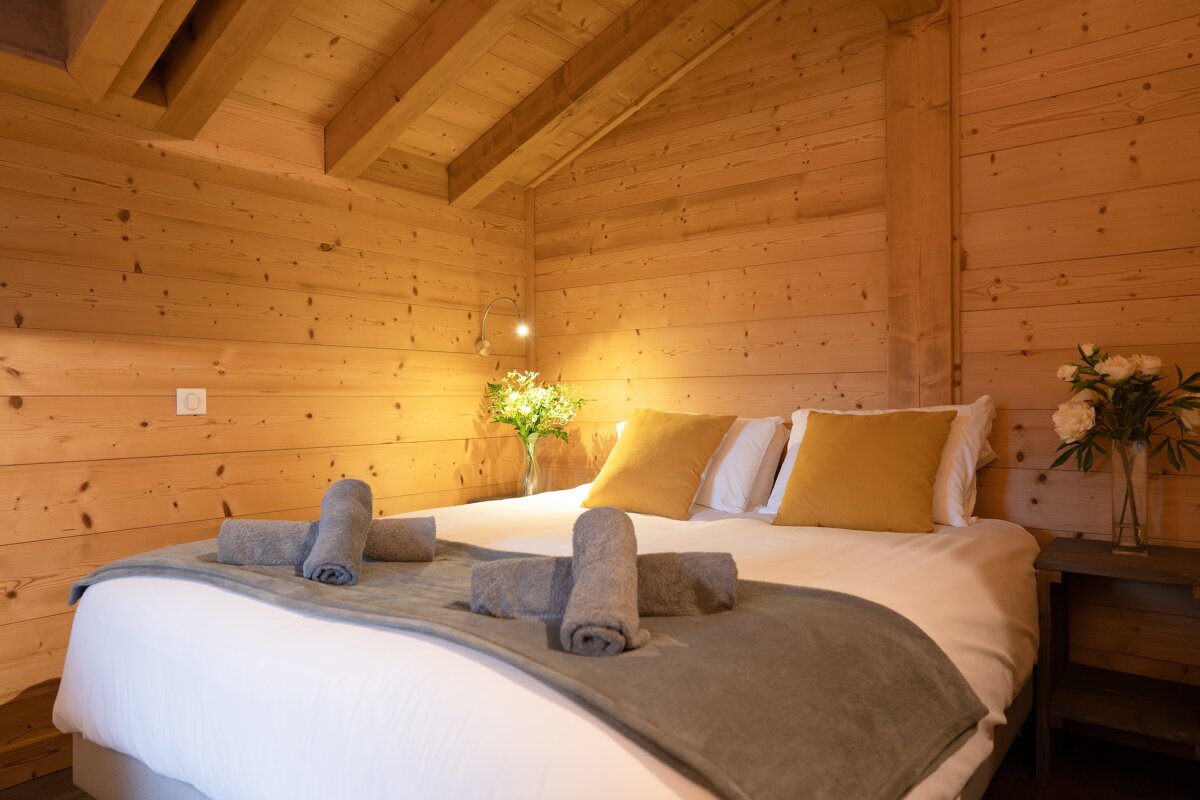 A warm, wood-paneled bedroom features a large bed with white linens, yellow pillows, a grey throw, and rolled towels. Flowers adorn nightstands.