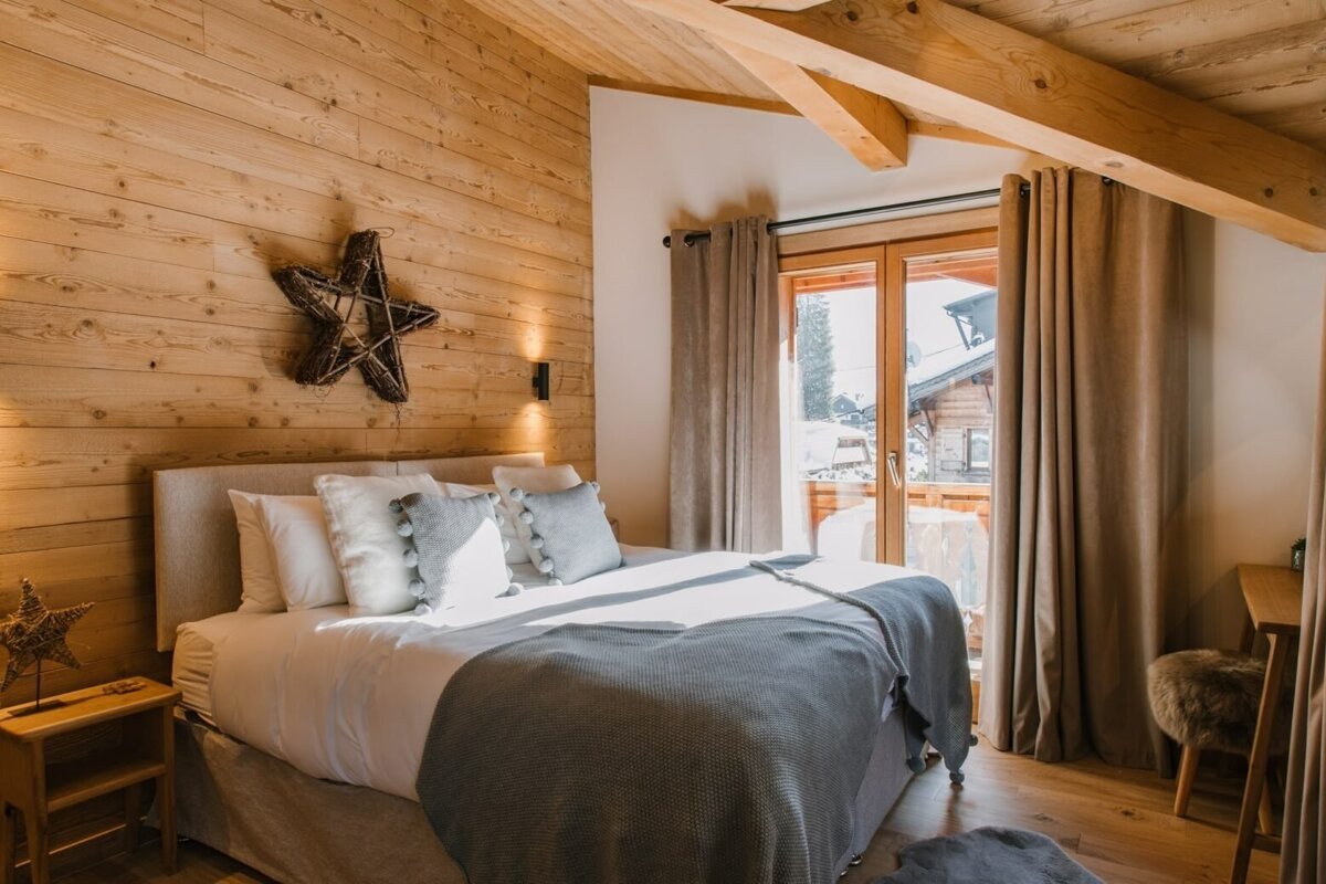 A cozy, rustic bedroom with wood-paneled walls, exposed beams, and a bed with gray bedding. Sunlight streams through a balcony door showing a snowy view.