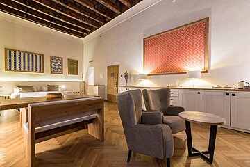 A chic hotel suite featuring a wooden beamed ceiling, herringbone floor, large bed, cozy armchairs, and modern art on white walls.