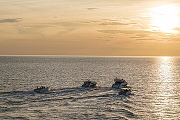Four boats cruise on a golden ocean at sunset, leaving wakes as the sun reflects brightly on the water under a warm sky.