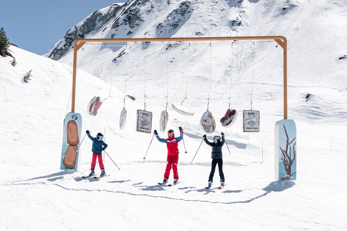 Three happy skiers pose under a cowboy-themed arch with targets on a sunny, snowy mountain slope, framed by a vast mountain.