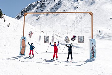 Three happy skiers pose under a cowboy-themed arch with targets on a sunny, snowy mountain slope, framed by a vast mountain.