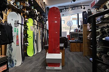 Inside a snowboard shop, a tall red snowboard stands prominently. Rows of various snowboards, boots, and gear line the walls.