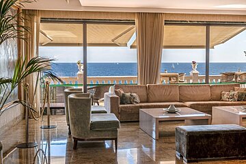 Elegant room with expansive sea views from large windows. Features comfortable sofas, chairs, marble floors, and distant sailboats on the ocean.