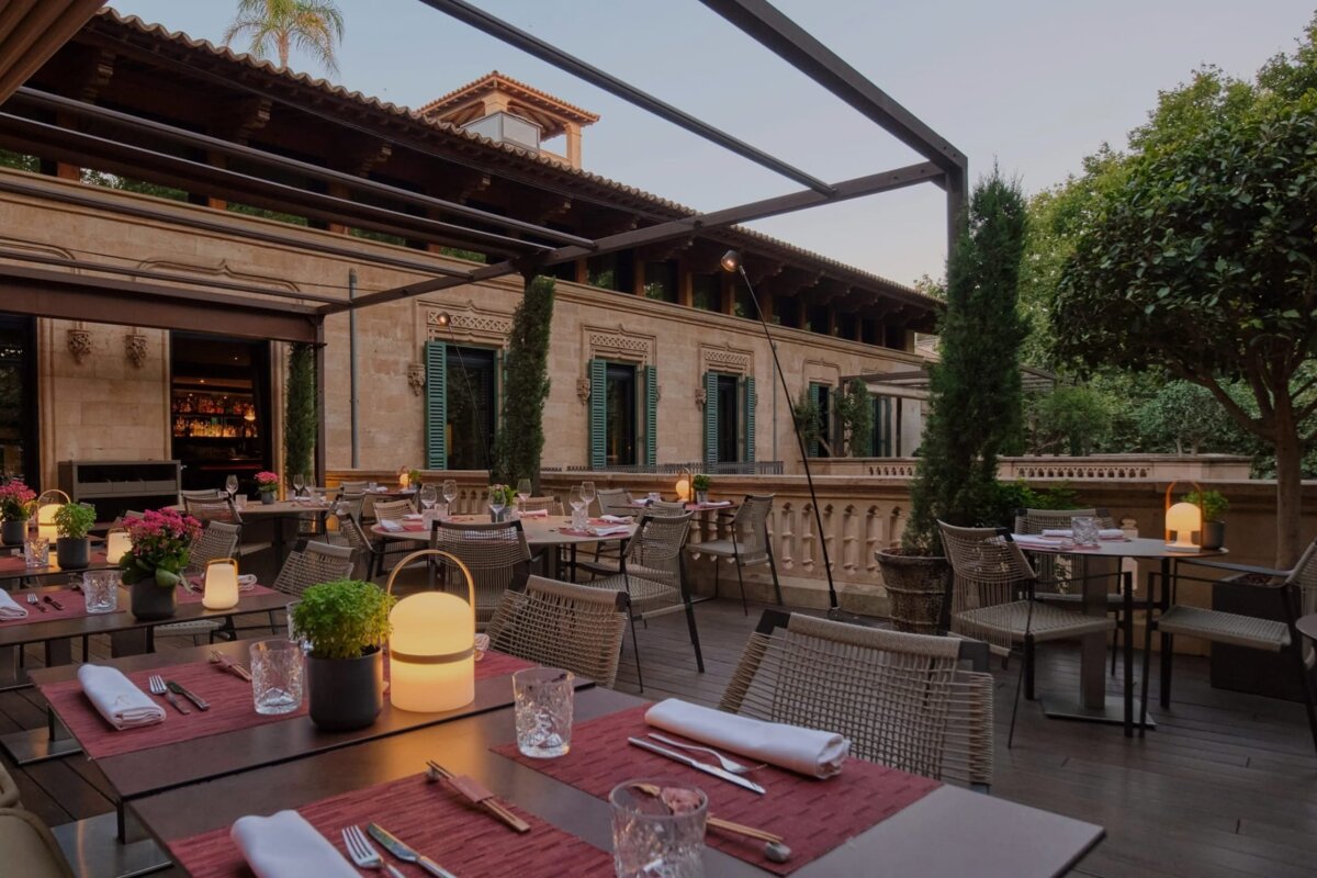 An elegant outdoor restaurant terrace with set tables, glowing lanterns, and a historic stone building in the background, viewed at dusk.