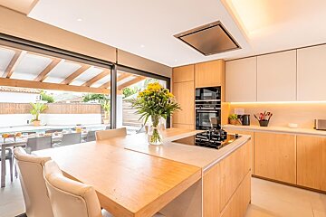 Bright, modern kitchen with light wood cabinetry, island with dining table, and glass doors leading to an outdoor pool and patio dining.