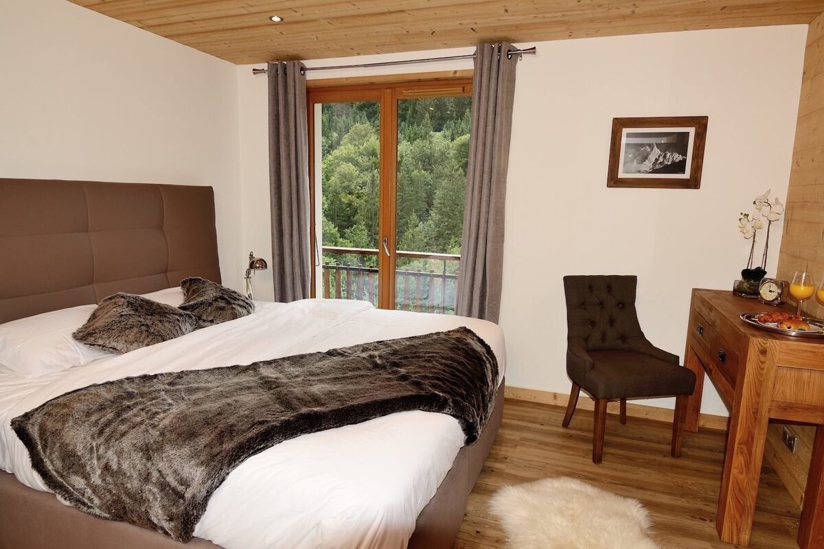 A cozy bedroom features a large bed with faux fur, a balcony overlooking a lush forest, and a wooden dresser with a breakfast tray.