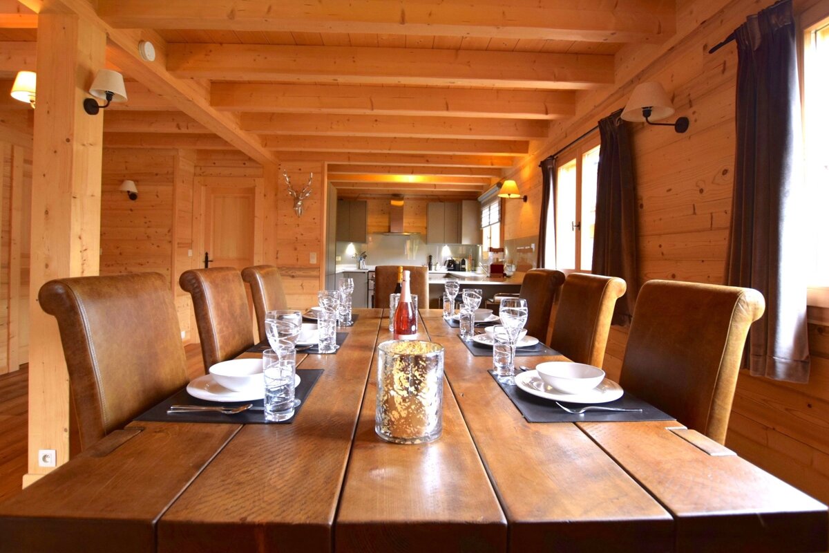 A warm, all-wood chalet dining room with a long table set for a meal, flanked by brown leather chairs, and a modern kitchen visible in the background.
