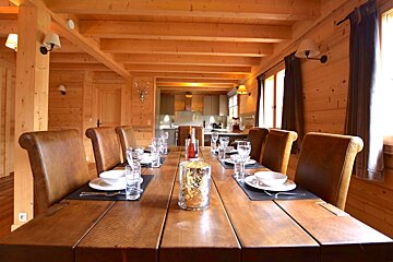 A warm, all-wood chalet dining room with a long table set for a meal, flanked by brown leather chairs, and a modern kitchen visible in the background.