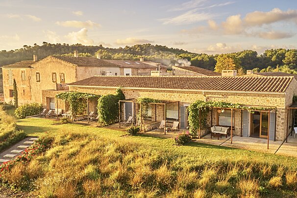 Picturesque stone hotel with terracotta roofs and vine-covered patios, nestled in golden grass and green hills under a warm, cloudy sky.