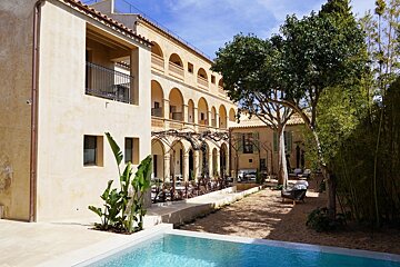 Sunny courtyard with a clear blue pool, a light-colored arched hotel building, trees, and bamboo under a blue sky.
