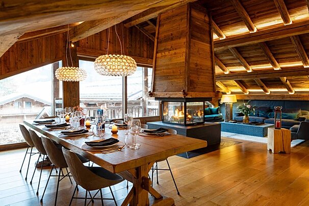 Luxury rustic chalet with a set wooden dining table, a modern glass fireplace, and a cozy blue living space. Exposed beams and large windows add charm.