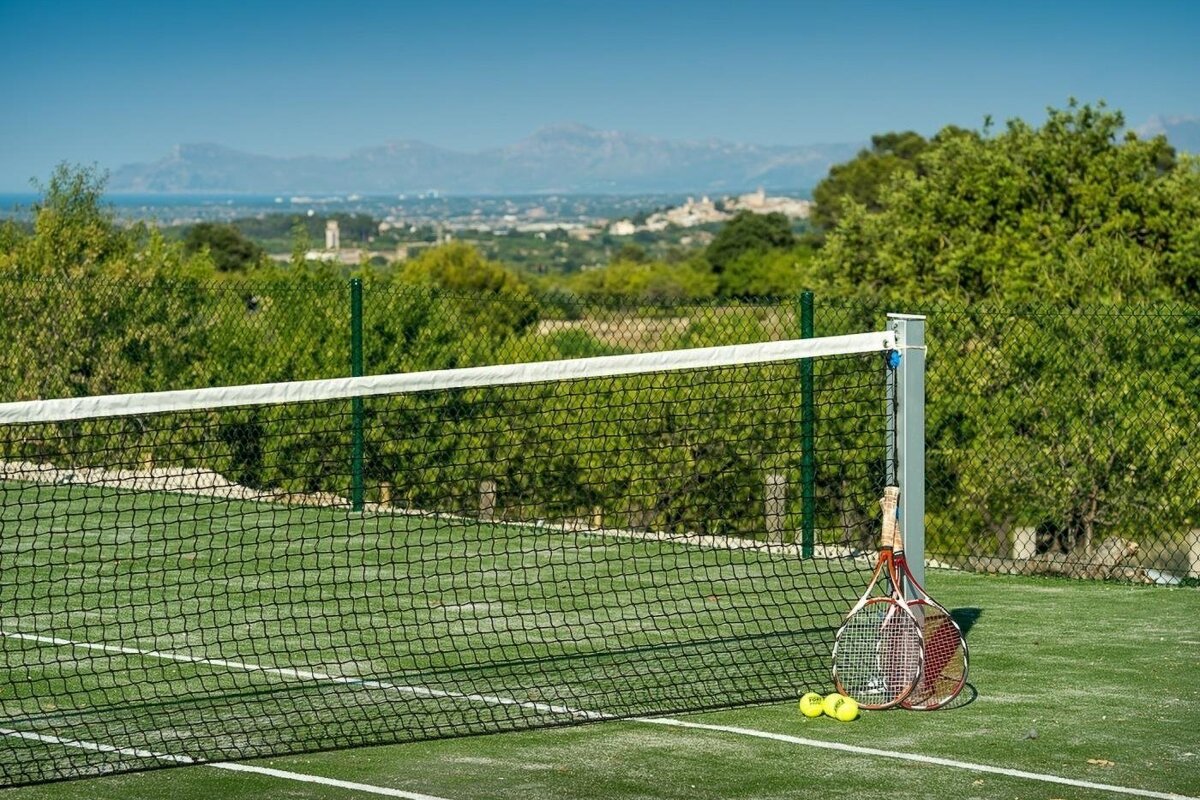 A green tennis court with racquets, balls, and a net, overlooking a scenic landscape with a town, mountains, and sea under a blue sky.