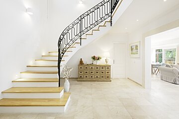 A bright, spacious hallway featuring a curved wooden staircase with ornate black iron railings, tile floors, and a wooden chest.