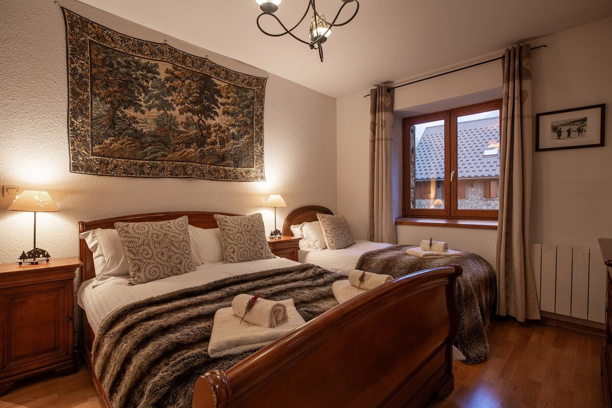 Cozy bedroom with a queen bed, twin bed, wooden furniture, and faux fur throws. A tapestry hangs above the main bed, and a window overlooks rooftops.