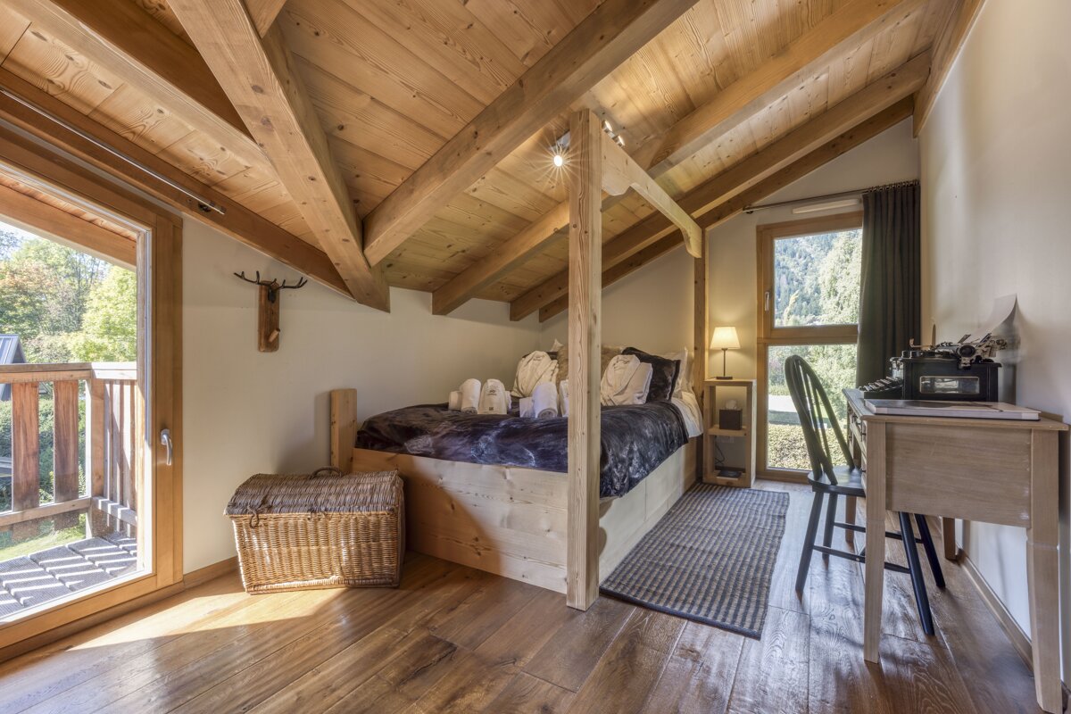A cozy, wood-paneled attic bedroom features a bed with towels, a desk with a typewriter, and a balcony overlooking nature. Rustic charm and natural light.