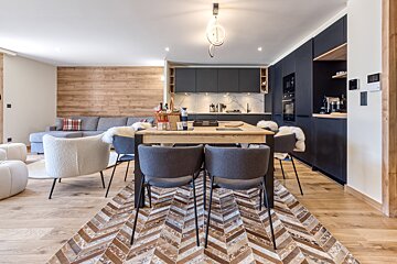 A sleek open-plan living, dining, and kitchen space. Features include dark cabinetry, light wood floors, a chevron rug, and a rustic wood accent wall.