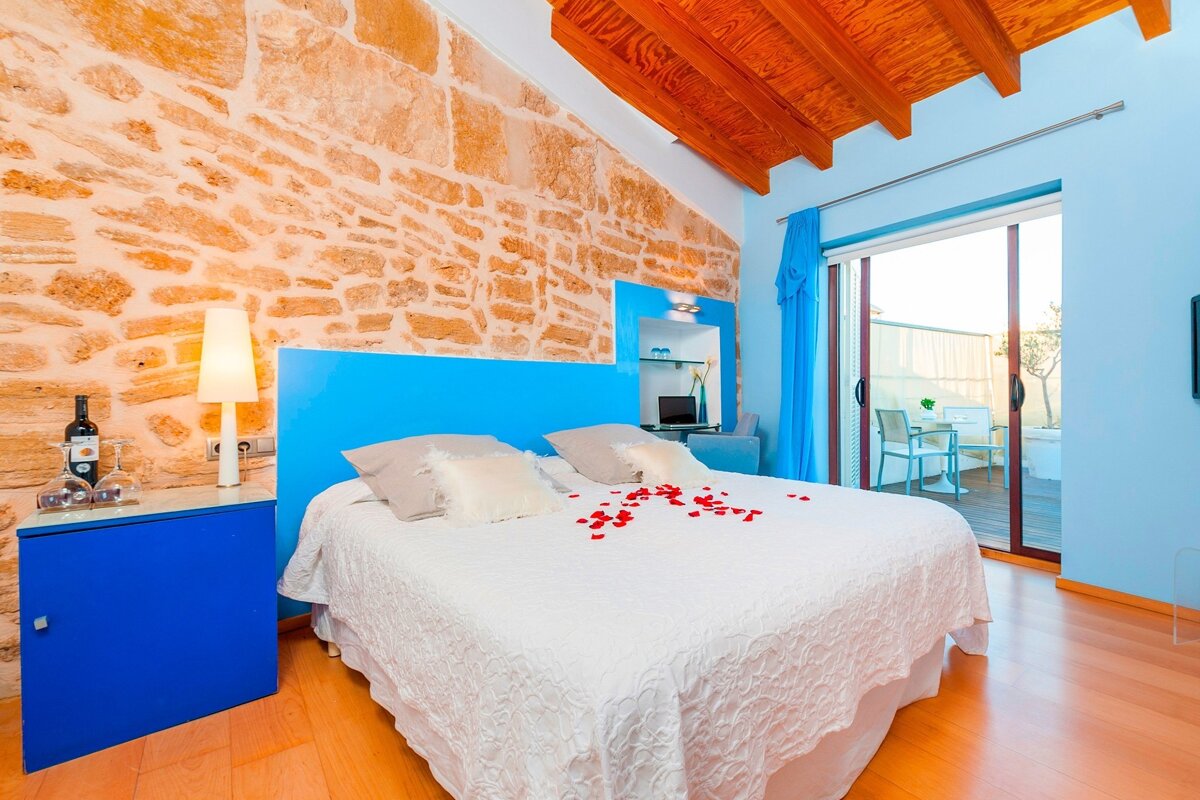 Rustic bedroom with a stone wall, blue accents, rose petals on the bed, wooden ceiling, and a balcony view.