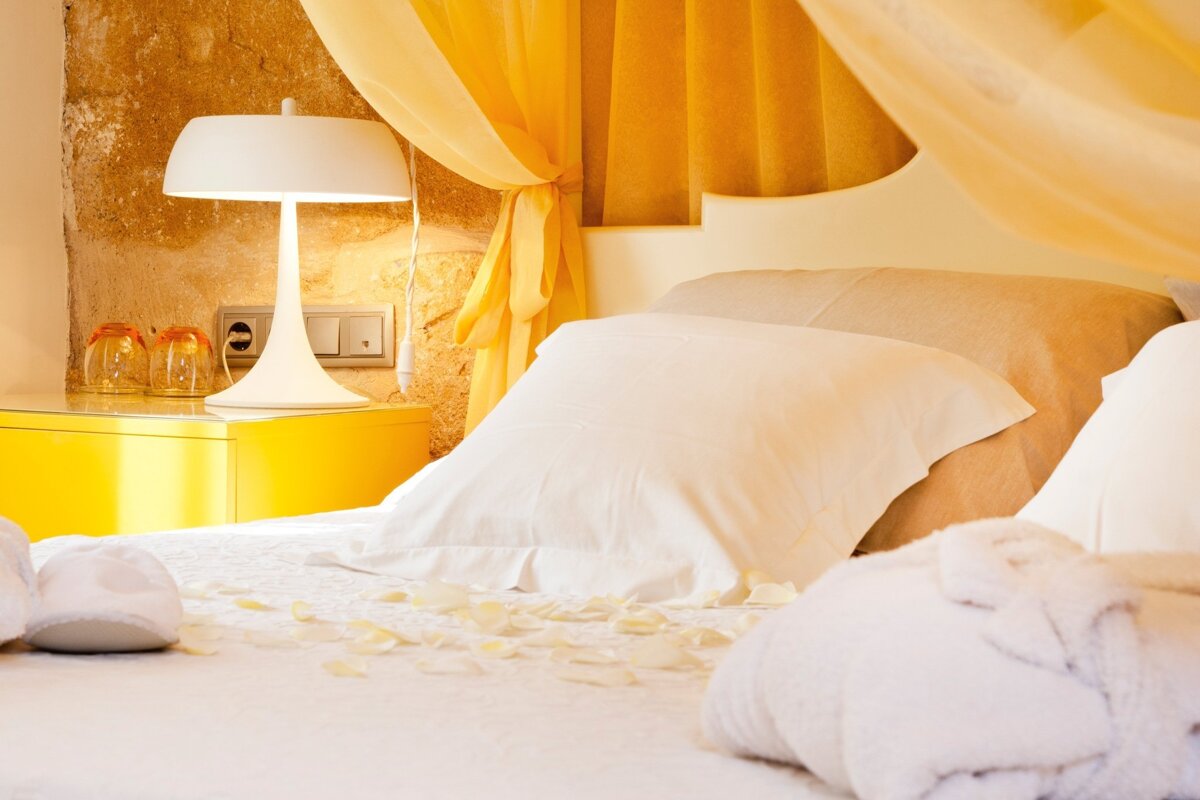 A warm, inviting bedroom features a white bed with rose petals, a bathrobe, and slippers. Yellow curtains and a nightstand with a lamp add color.