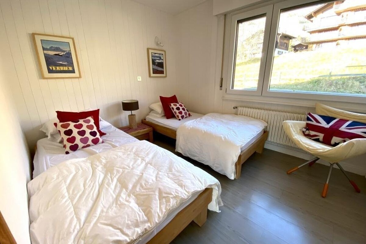Bright twin bedroom with white paneled walls, wood floor, and a window to a green hill. Beds have white duvets and colorful pillows.