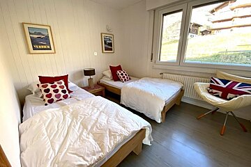 Bright twin bedroom with white paneled walls, wood floor, and a window to a green hill. Beds have white duvets and colorful pillows.