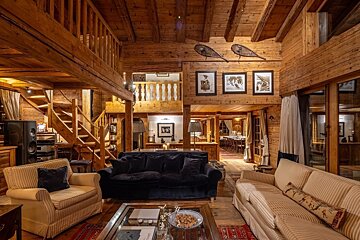 A cozy, multi-level wooden chalet living room with plush sofas, rustic decor including snowshoes, and a visible dining area, exuding warmth and comfort.
