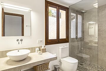 A modern bathroom featuring a concrete vanity with a vessel sink, a wooden window, a toilet, and a glass-enclosed shower with pebble flooring.
