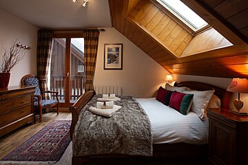 A cozy attic bedroom features a double bed with a furry throw, warm lighting, and a slanted wooden ceiling with a skylight. A balcony door and rustic furniture complete the inviting space.