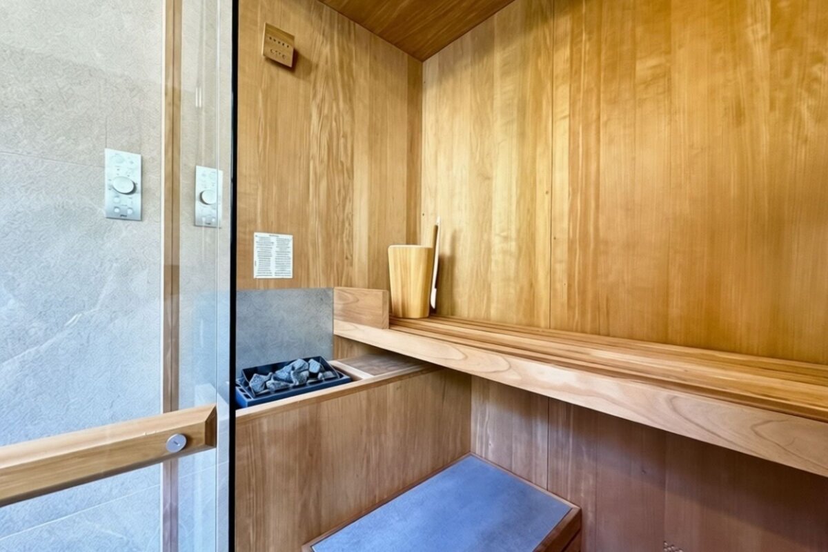 A modern wooden sauna interior with a heater, bucket, ladle, and multi-tiered bench. A glass door separates it from a tiled shower area with controls.