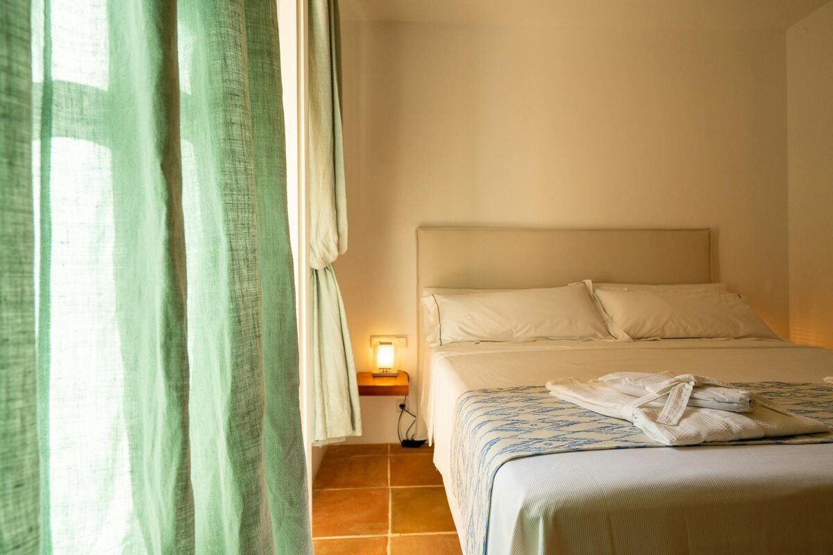 Bright bedroom with a white bed, patterned blue and white runner, and a white bathrobe. Light green curtains frame a window next to a warm lamp.
