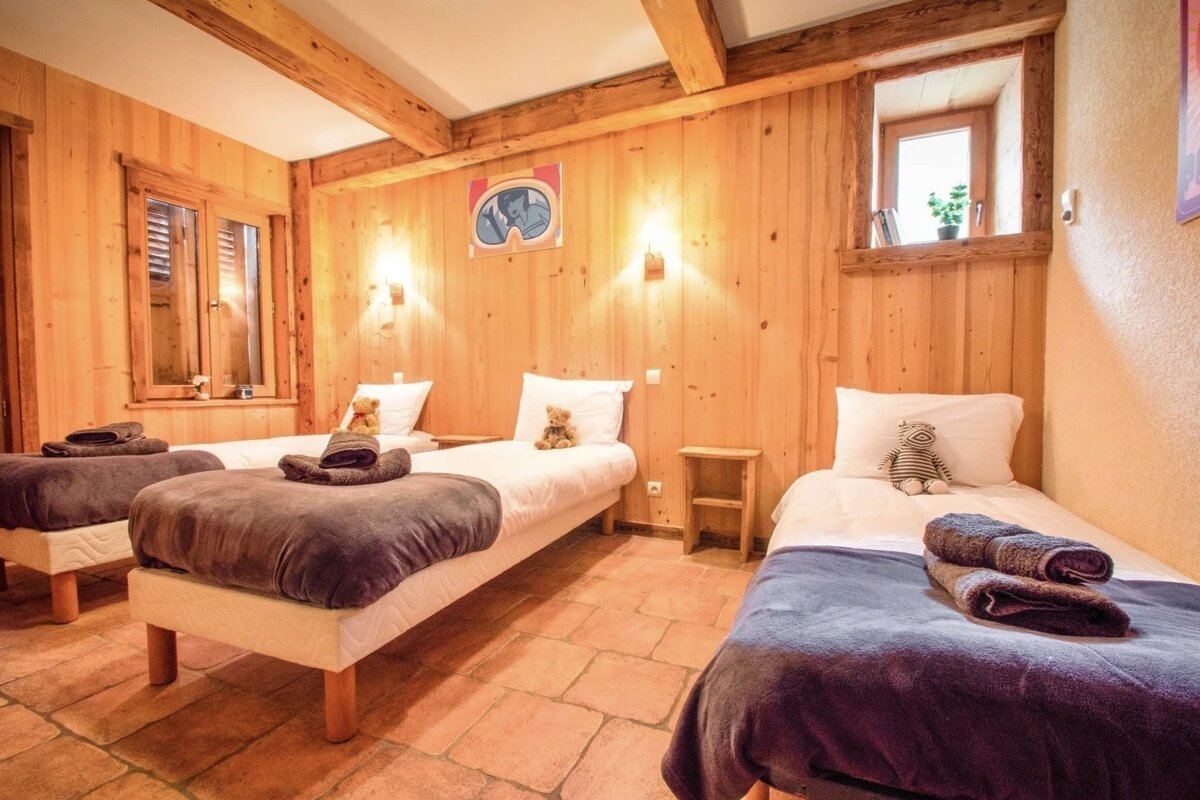A rustic, wood-paneled bedroom features three single beds with white linens and stuffed animals, exposed beams, and a terracotta tile floor.