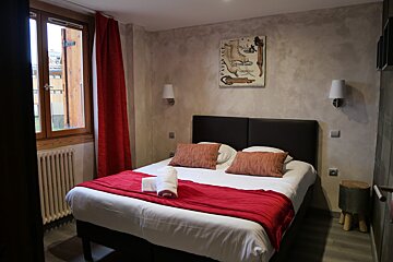 A cozy bedroom features a double bed with white bedding, red throw and curtains, textured walls, a window, wall art, and a small stool.