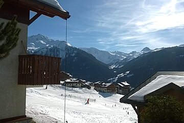 Chalet and balcony, snow covered roof overlooking a vallandry piste with a couple of buildings in the background, with a backdrop of snowy mountains and a blue sky