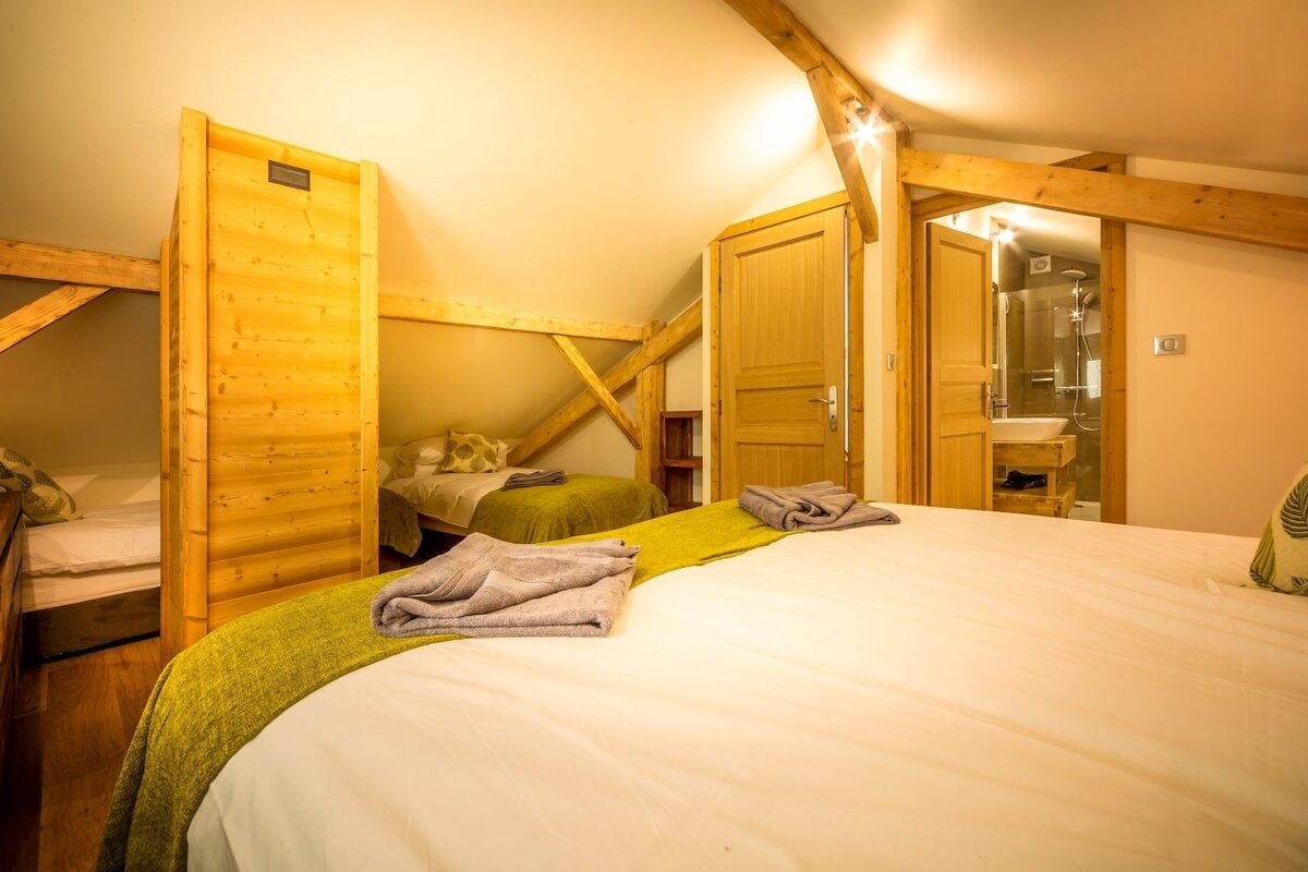 A warm, attic-style bedroom with exposed wooden beams, a main bed, two smaller beds, and a visible en-suite bathroom. Cozy, chalet ambiance.
