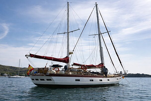 A large white and red sailboat with two masts and furled red sails floats on calm blue water. A Spanish flag flies from its bow.